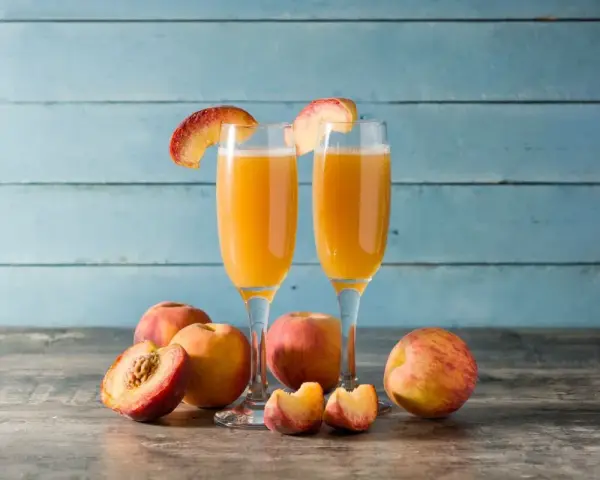 Dwa kieliszki z orzeźwiającym drinkiem na bazie likieru brzoskwiniowego, ozdobione plasterkami brzoskwini. Obok świeże owoce.