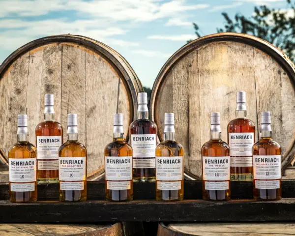 Butelki whisky BenRiach stoją przed beczkami, sugerując skąd pochodzi whisky.