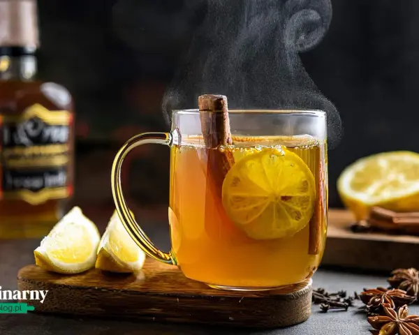 Gorąca herbata z whisky, cytryną i cynamonem, parująca w szklance. Obok leżą cytryny i przyprawy.