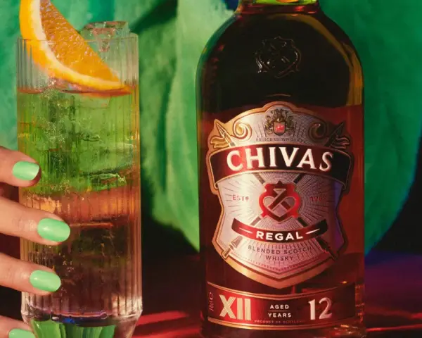 Chivas Regal 12 z czym pić? Orzeźwiający drink z plasterkiem pomarańczy i lodem obok butelki Chivas Regal 12.