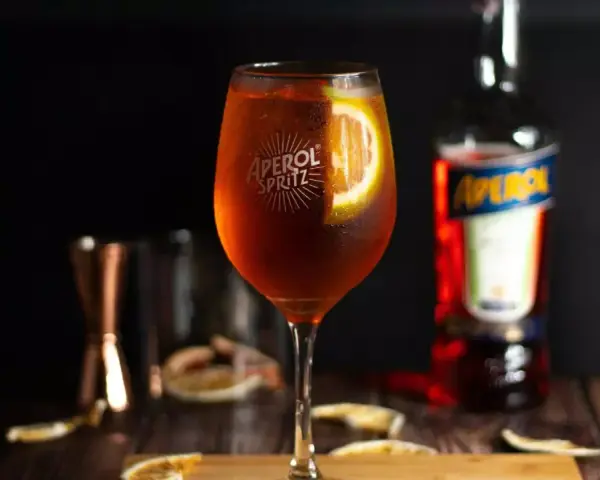Orzeźwiający Aperol ze Sprite, udekorowany plasterkiem pomarańczy, na drewnianym blacie. Obok butelka Aperolu i inne akcesoria barmańskie.