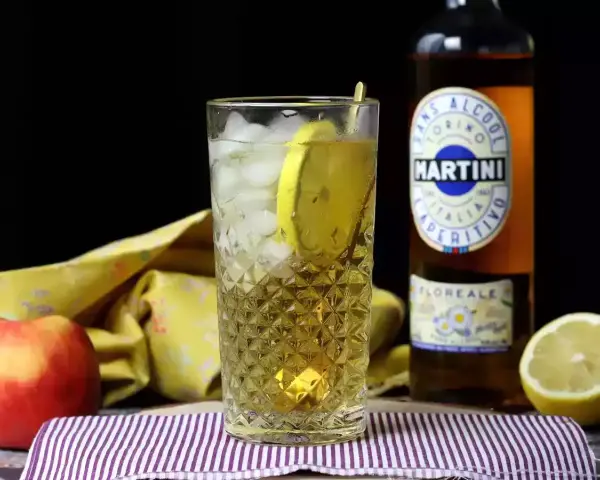Orzeźwiający drink z Martini i lodem, ozdobiony plasterkiem cytryny. Obok jabłko i butelka Martini.