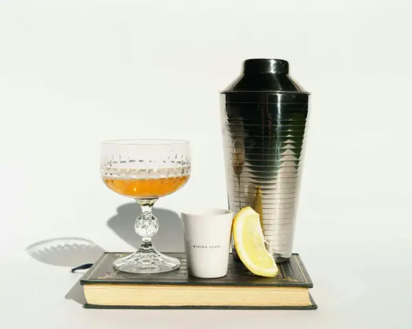 Elegancki kieliszek z bursztynowym trunkiem, shaker, plasterek cytryny i biały kubeczek z napisem "MIXING GLASS" – wszystko to sugeruje, z czym pić whisky, by stworzyć wyrafinowany koktajl.