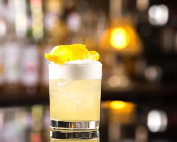 Orzeźwiający drink z whisky i miodem, ozdobiony skórką cytryny, na tle rozmytego baru.
