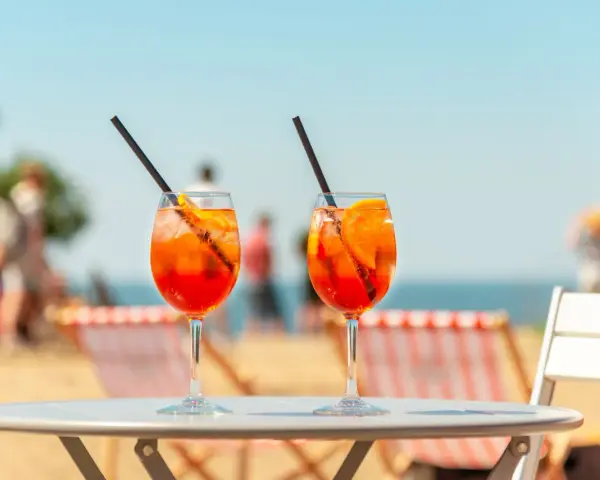 Dwa orzeźwiające drinki aperol z sokiem pomarańczowym, z lodem i plasterkiem pomarańczy, na tle plaży i błękitnego nieba. Idealne na wakacyjny relaks.