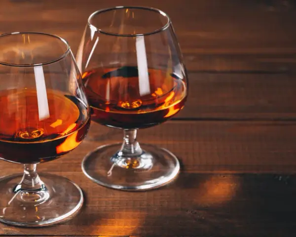 Dwa kieliszki z bursztynowym płynem, być może amerykańska whisky z kukurydzy, stoją na drewnianym stole.
