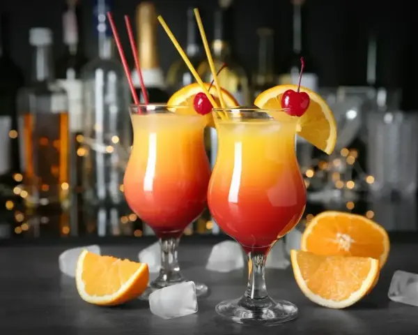 Dwa drinki z pomarańczą i wiśnią, ozdobione słomkami, na tle butelek i światełek.