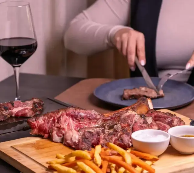 Idealne wino do dziczyzny: kieliszek czerwonego wina obok soczystego steka i frytek. Ktoś kroi mięso na talerzu.