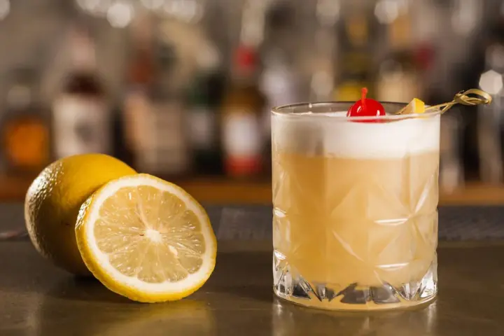 Orzeźwiający drink z amaretto, ozdobiony wisienką i plasterkiem cytryny, obok świeżych cytryn na barze.