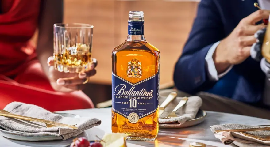 Elegancka kolacja z whisky i piwem w tle. Butelka Ballantine's 10 Years Old na stole, obok kieliszek z bursztynowym trunkiem.