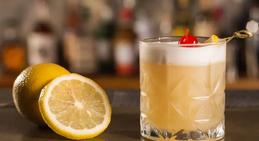 Orzeźwiający drink z amaretto, ozdobiony wisienką i plasterkiem cytryny, obok świeżych cytryn na barze.