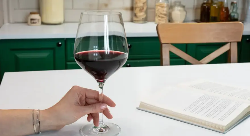 Dłoń elegancko trzyma kieliszek z czerwonym winem, pokazując, jak trzymać kieliszek do wina. Obok leży otwarta książka.