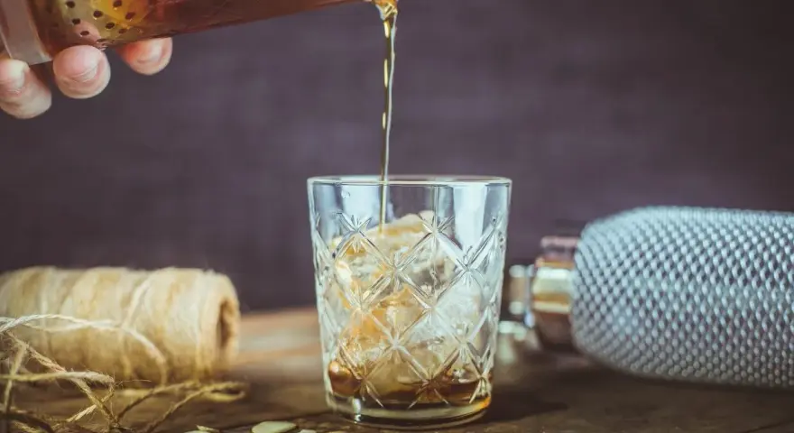Nalewanie drinka z amaretto i wódką do szklanki z lodem. Na stole płatki migdałów i sznurek.