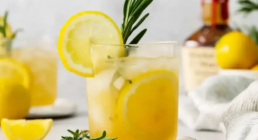 Szklanka z drinkiem z cytryną i rozmarynem, w tle butelka alkoholu i cytryny.