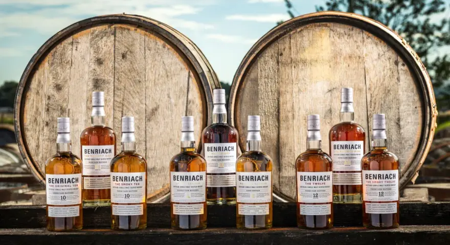 Butelki whisky BenRiach stoją przed beczkami, sugerując skąd pochodzi whisky.