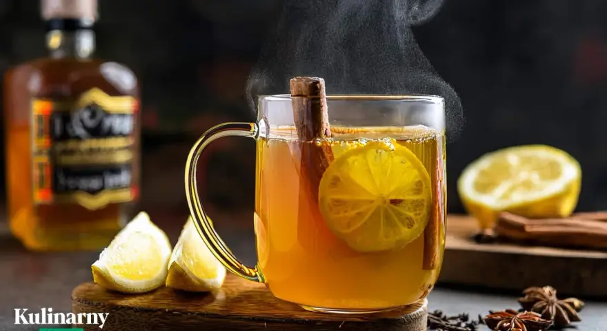 Gorąca herbata z whisky, cytryną i cynamonem, parująca w szklance. Obok leżą cytryny i przyprawy.