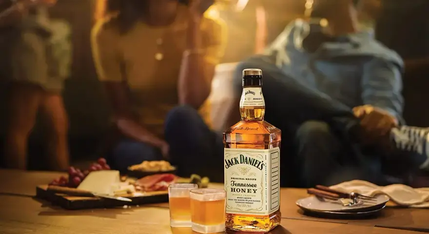 Jack Daniel's Honey z czym pić? Idealnie smakuje solo, z lodem lub jako dodatek do drinków. Na stole ser, winogrona i przekąski.