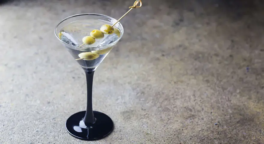 Elegancki martini z ginem, ozdobione trzema oliwkami na wykałaczce, w czarnej kieliszku na kamiennym tle.