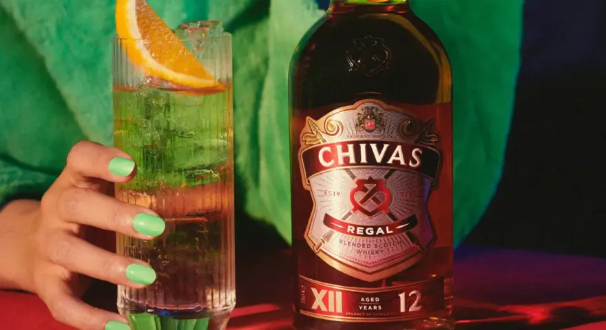 Chivas Regal 12 z czym pić? Orzeźwiający drink z plasterkiem pomarańczy i lodem obok butelki Chivas Regal 12.