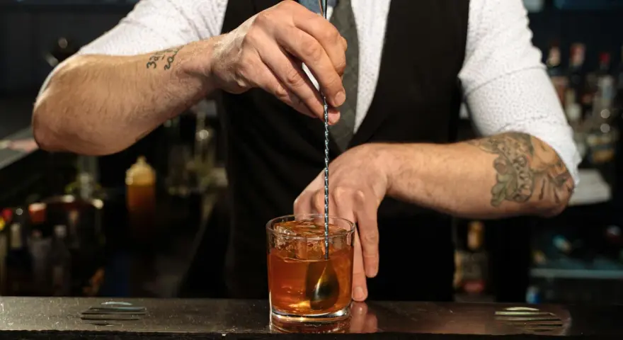 Barman miesza kostki lodu z bursztynowym płynem w szklance. Idealny **whisky z tonikiem** gotowy do podania.