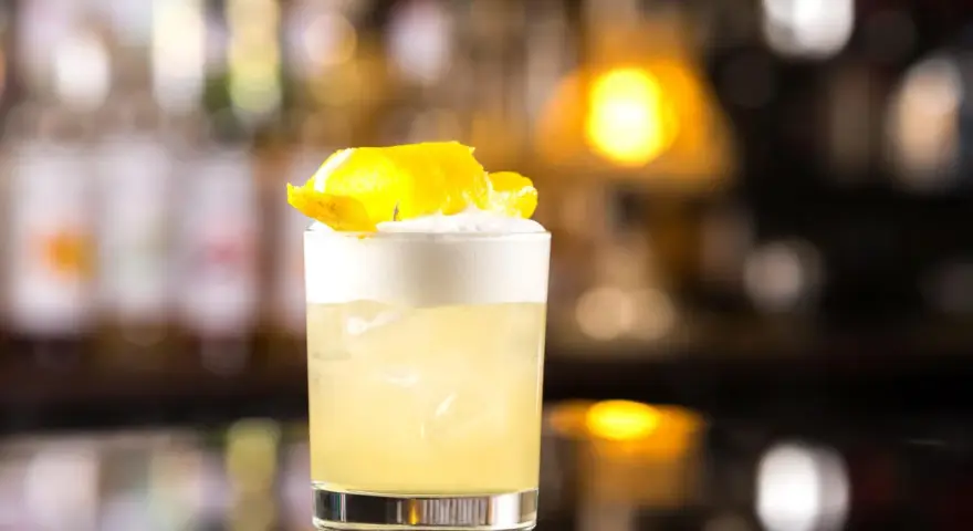 Orzeźwiający drink z whisky i miodem, ozdobiony skórką cytryny, na tle rozmytego baru.