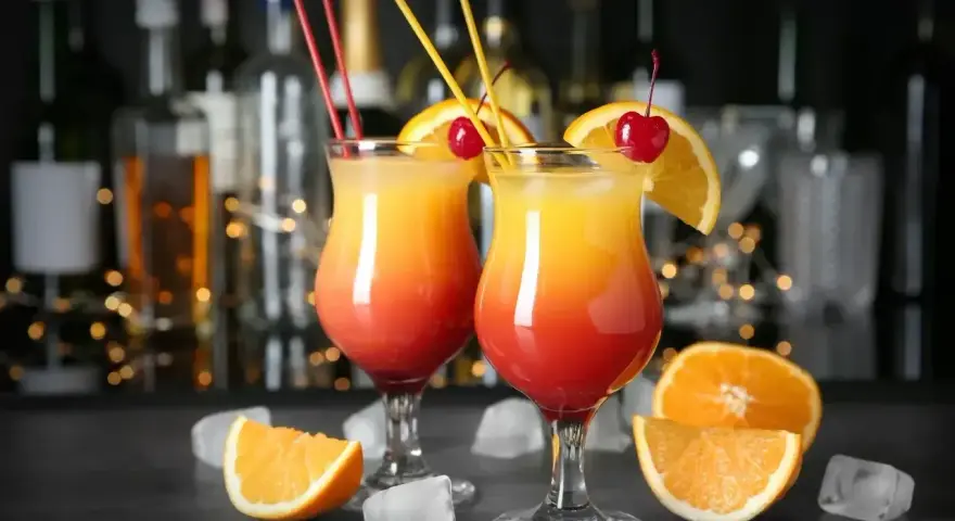 Dwa drinki z pomarańczą i wiśnią, ozdobione słomkami, na tle butelek i światełek.