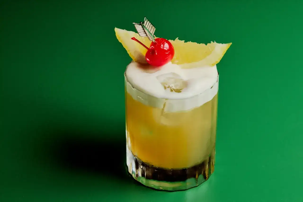 Amaretto sour przepis: drink z pianką, wiśnią i cytryną na zielonym tle.
