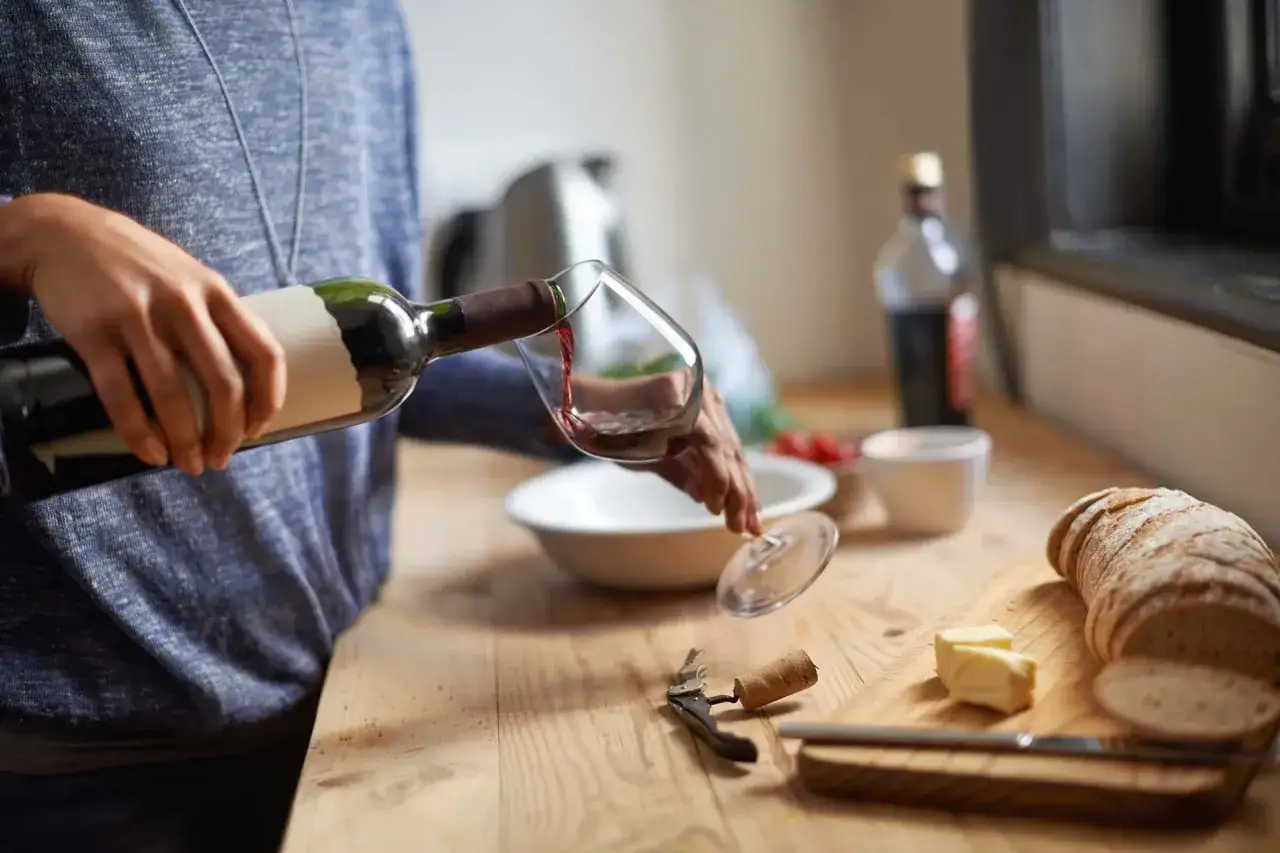 Ręka nalewa czerwone wino do kieliszka. Na drewnianym blacie leży chleb, masło i korkociąg.
