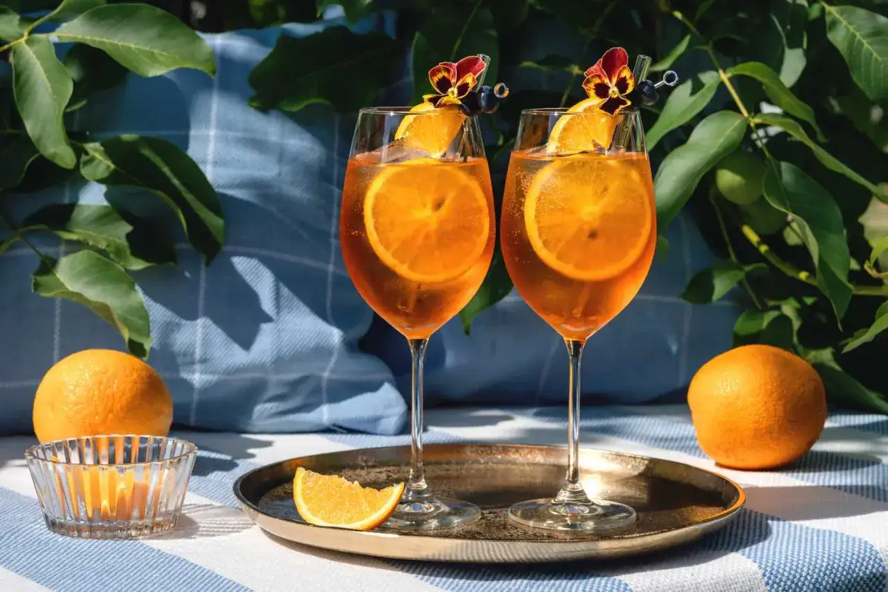 Dwa orzeźwiające aperole z sokiem pomarańczowym, ozdobione plastrami pomarańczy i kwiatami, gotowe do degustacji w letnim słońcu.
