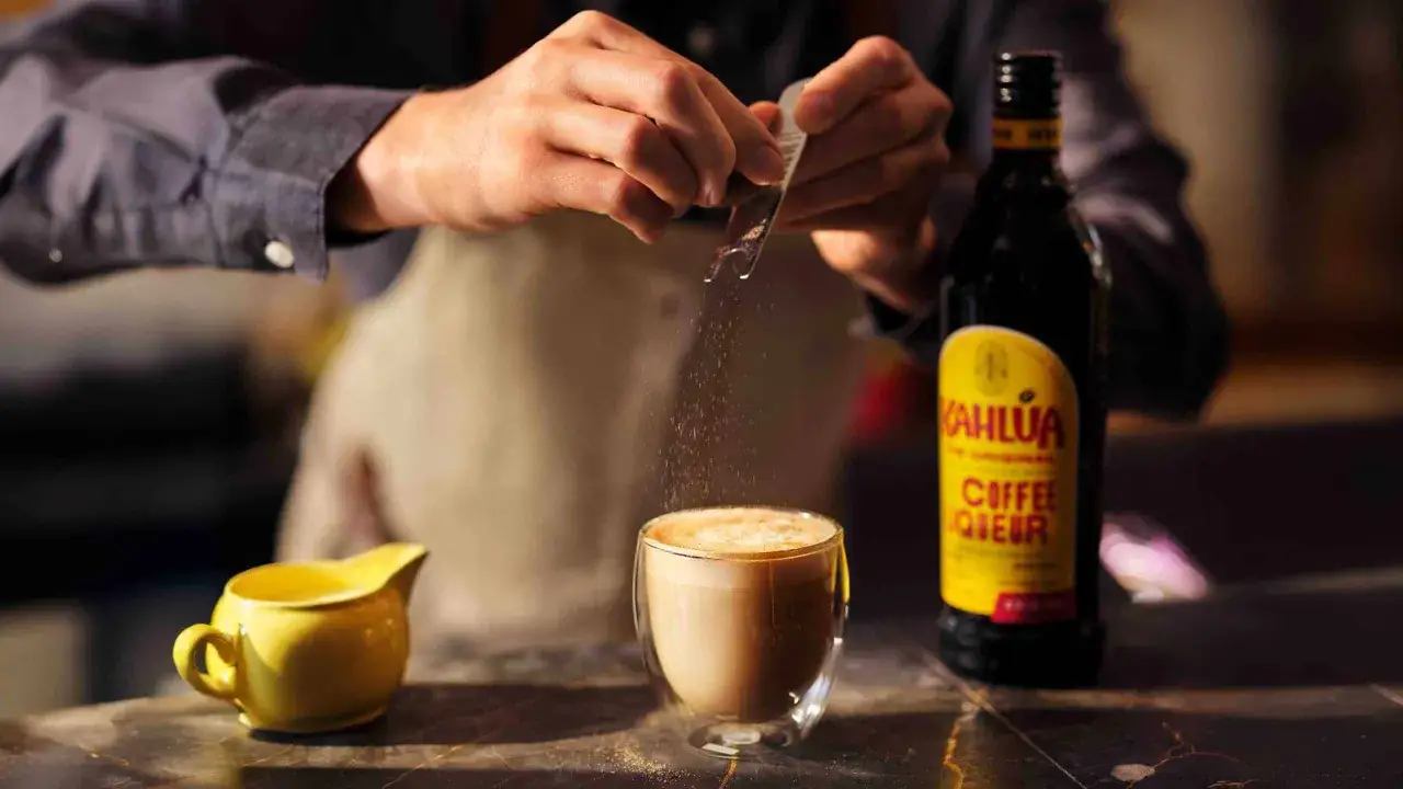 Barista dodaje cukier do kawy z Kahl&uacute;a. Butelka Kahl&uacute;a i mały dzbanuszek stoją obok.