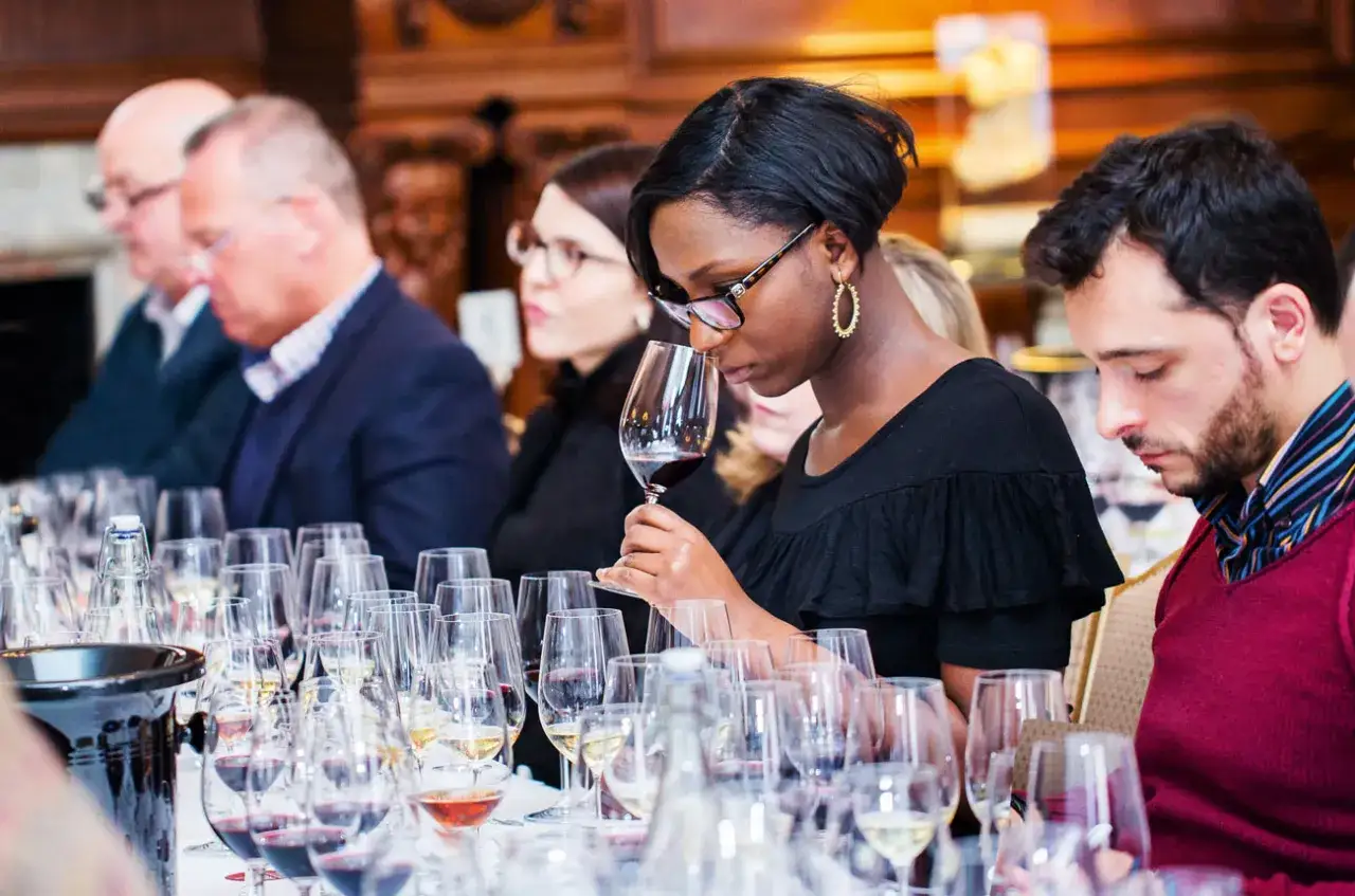 Kobieta wącha wino, ucząc się, jak degustować wino. Wok&oacute;ł stołu pełnego kieliszk&oacute;w z winem siedzą inni uczestnicy.