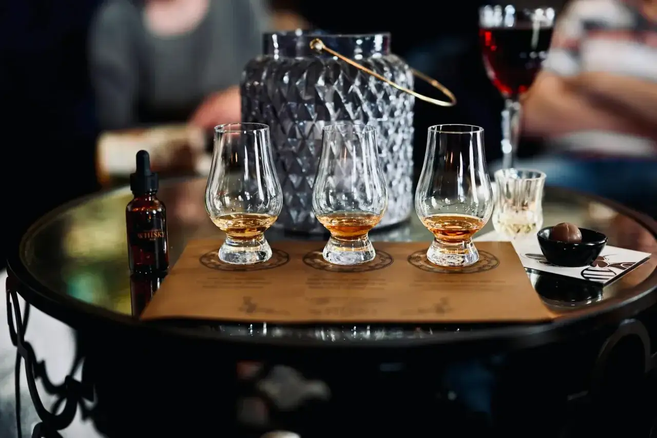 Trzy kieliszki z whisky, buteleczka z kroplomierzem i czekoladka na stole. Idealne do degustacji whisky online.
