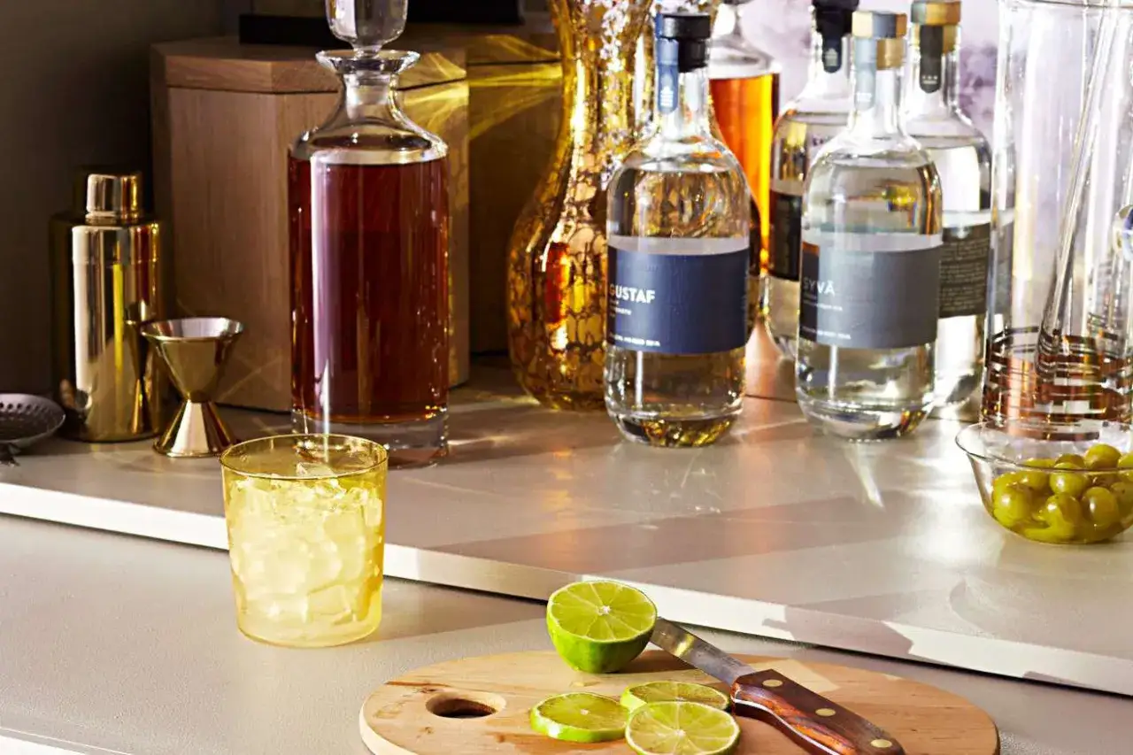 Na blacie stoją podstawowe alkohole do drink&oacute;w: whisky, w&oacute;dka i gin. Obok szklanka z lodem, limonka i oliwki.