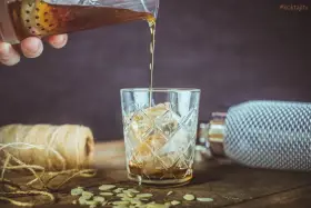 Nalewanie drinka z amaretto i wódką do szklanki z lodem. Na stole płatki migdałów i sznurek.