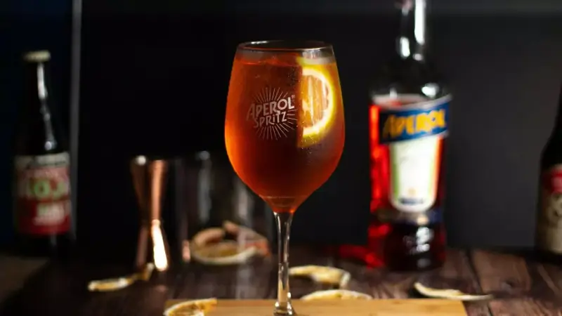Orzeźwiający Aperol ze Sprite, udekorowany plasterkiem pomarańczy, na drewnianym blacie. Obok butelka Aperolu i inne akcesoria barmańskie.
