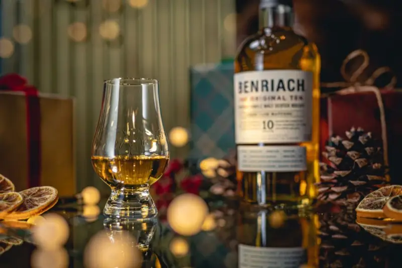 Szklanka z bursztynowym single malt whisky Benriach 10, obok butelki i świątecznych dekoracji.