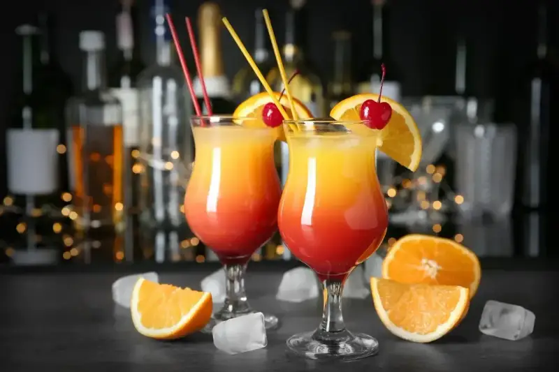 Dwa drinki z pomarańczą i wiśnią, ozdobione słomkami, na tle butelek i światełek.